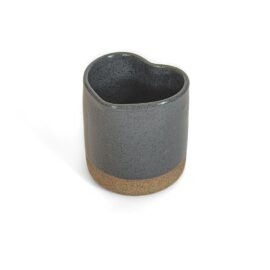 Copo Coração da Esmo Cerâmica na cor Cinza, visto em um ângulo oblíquo sobre fundo branco. A peça de cerâmica artesanal possui formato ergonômico de coração. O corpo superior apresenta acabamento esmaltado escuro e brilhante com ricos pontilhados texturizados, criando um encontro de texturas direto com a base inferior exposta em cerâmica crua de tom terroso claro.