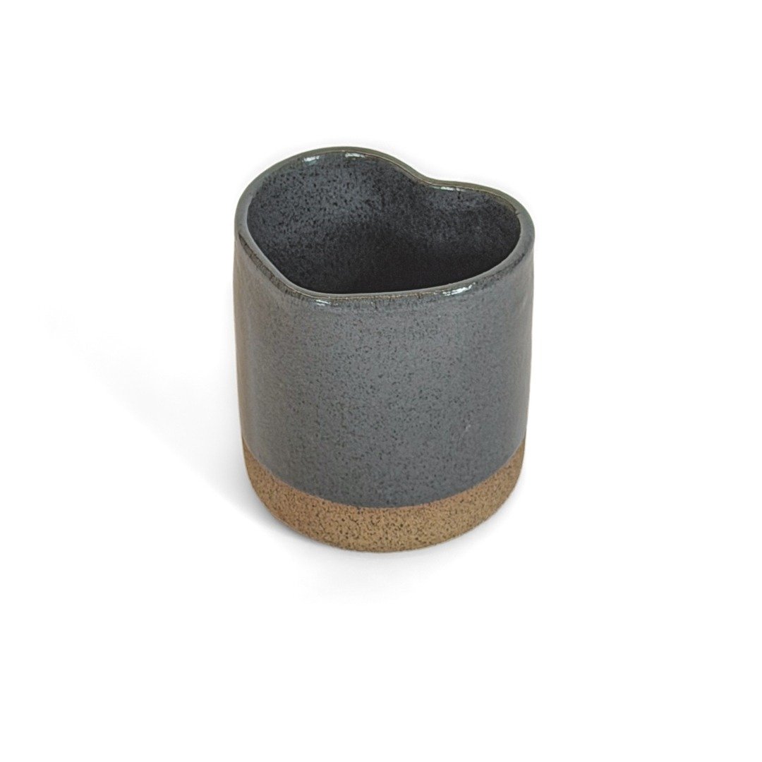 Copo Coração da Esmo Cerâmica na cor Cinza, visto em um ângulo oblíquo sobre fundo branco. A peça de cerâmica artesanal possui formato ergonômico de coração. O corpo superior apresenta acabamento esmaltado escuro e brilhante com ricos pontilhados texturizados, criando um encontro de texturas direto com a base inferior exposta em cerâmica crua de tom terroso claro.