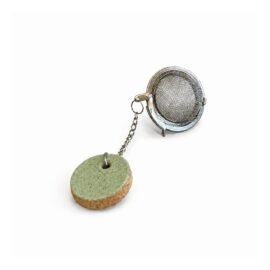 O infusor de chá com Tag Redonda em Cerâmica Artesanal da Esmo (3cm) ganha frescor na edição Pistache. Uma peça utilitária que funciona como contrapeso estratégico na xícara, destacando a dualidade visual entre o esmalte verde suave e a rusticidade natural da matéria exposta na base.