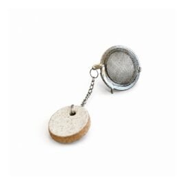 A Tag Redonda com Infusor de Chá da Esmo em Cerâmica Artesanal fotografada sobre fundo branco limpo. A peça utilitária exibe um infusor esférico metálico de malha fina em aço, conectado por uma delicada corrente a uma pequena tag circular em cerâmica artesanal na cor Off-White. A iluminação clara destaca a textura orgânica pontilhada e a superfície mate da face superior da tag, além do contraste tátil de sua lateral em cerâmica natural aparente. A composição reflete precisão geométrica e pureza formal, com sombras suaves que projetam a silhueta do objeto e enfatizam a sua clareza visual e funcionalidade.