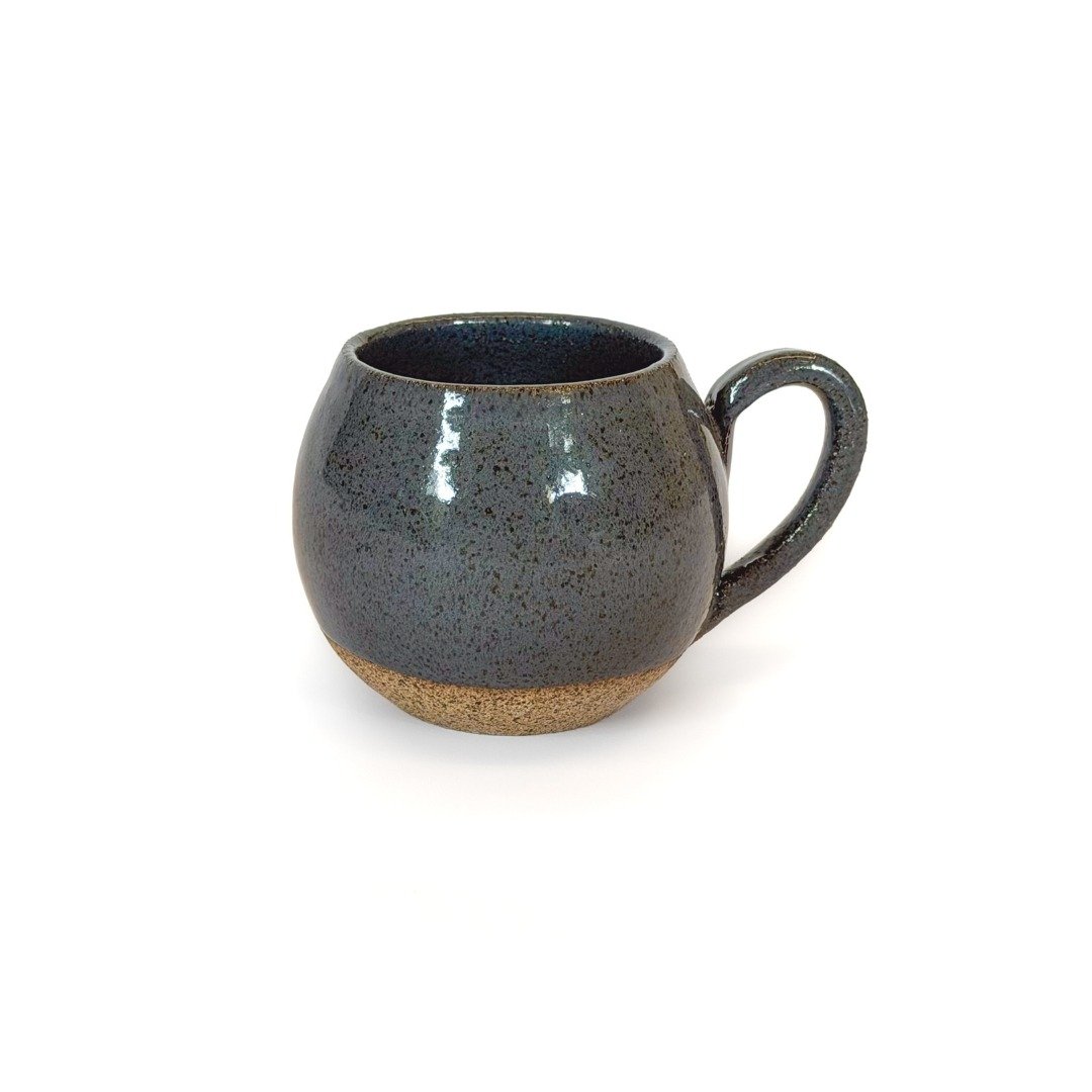 Xícara Esmo Longo de cerâmica com formato arredondado. A parte superior possui um esmalte cinza escuro brilhante com micropartículas e textura salpicada que reflete a luz, contrastando diretamente com a faixa inferior da base em argila bruta exposta de tom terroso texturizado. Fundo branco.