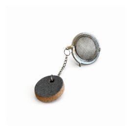 Infusor de chá metálico em malha fina, conectado por uma corrente a uma tag redonda de cerâmica artesanal com face cinza-escura texturizada e lateral de massa natural aparente, fotografado sobre fundo branco. Esmo Cerâmica