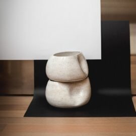 Composição com dois Centros de Mesa Alto Grande na cor Off-White de cerâmica empilhados, evidenciando o contraste da silhueta orgânica irregular e o acabamento salpicado suave contra um fundo geométrico e minimalista.