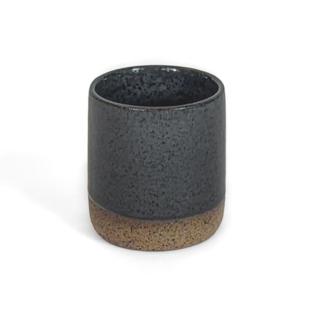 Copo Reto Grande em cerâmica artesanal na cor Cinza da marca Esmo Cerâmica. A peça possui formato cilíndrico e silhueta minimalista, com a parte superior finalizada em acabamento esmaltado com brilho escuro e textura pontilhada, enquanto a base exposta exibe o tom e a textura da cerâmica crua. Fotografia de produto com fundo branco.