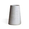 Vaso Cônico Grande na cor Off-White da Esmo. A peça decorativa em cerâmica artesanal possui silhueta geométrica de linhas retas, afunilando em direção ao topo. O acabamento mate revela uma textura orgânica com sutis pontilhados escuros, difundindo a luz suavemente pela superfície. Na base ampla, observa-se um fino filete de cerâmica rústica escura aparente, gerando um elegante contraste visual e projetando uma leve sombra à esquerda sobre um fundo claro.