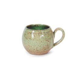 Xícara Esmo Longo em cerâmica na cor Pistache. Peça com silhueta arredondada e alça, apresentando forte contraste visual e tátil entre a parte superior esmaltada em verde suave, com acabamento vitrificado e pontilhado natural, e a base rústica em argila exposta.