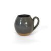Caneca Esmo de cerâmica autoral. A peça possui silhueta arredondada e é esmaltada no corpo e na alça com um tom cinza escuro brilhante coberto por micropintas pretas. A base exibe a argila exposta e texturizada sem esmalte em tom terroso natural.