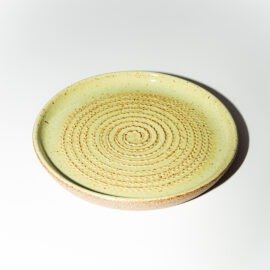 Prato Ralador de cerâmica da Esmo em perspectiva, na cor Matcha Latte. A peça exibe superfície esmaltada vitrificada com brilho em tom verde pálido e pontilhado escuro natural, ranhuras centrais em espiral e borda inferior com barro cru rústico exposto.