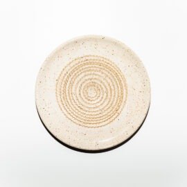 Vista superior do Prato Ralador na cor Off-White, produzido em cerâmica artesanal pela Esmo. A peça circular exibe uma superfície mate acetinada com uma textura sutil de minúsculas pontuações minerais integradas à matéria. No centro, sobressaem-se as ranhuras elevadas em padrão espiral contínuo, onde a luz incidente cria sombras suaves nos relevos cerâmicos, destacando a precisão geométrica e a funcionalidade do design projetado para ralar ingredientes.