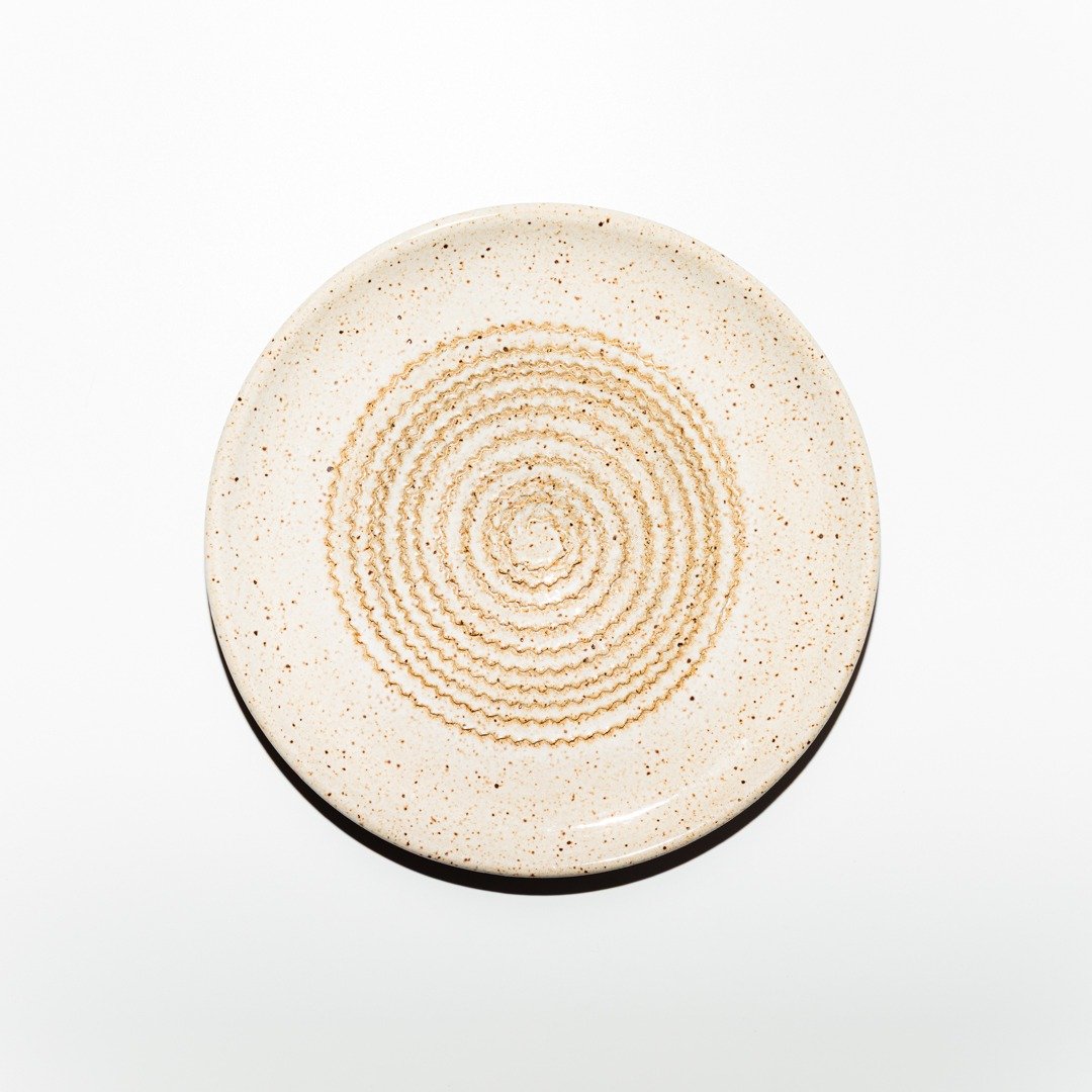 Vista superior do Prato Ralador na cor Off-White, produzido em cerâmica artesanal pela Esmo. A peça circular exibe uma superfície mate acetinada com uma textura sutil de minúsculas pontuações minerais integradas à matéria. No centro, sobressaem-se as ranhuras elevadas em padrão espiral contínuo, onde a luz incidente cria sombras suaves nos relevos cerâmicos, destacando a precisão geométrica e a funcionalidade do design projetado para ralar ingredientes.